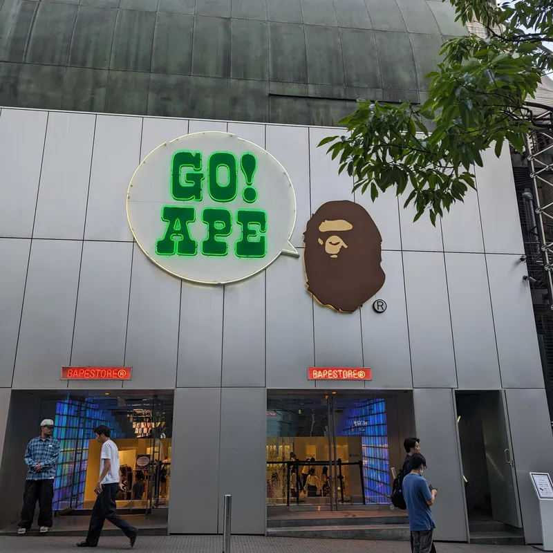 BAPE store exterior in Shibuya, Tokyo, Japan, with 'GO! APE' neon sign and ape head logo, pedestrians walking past.