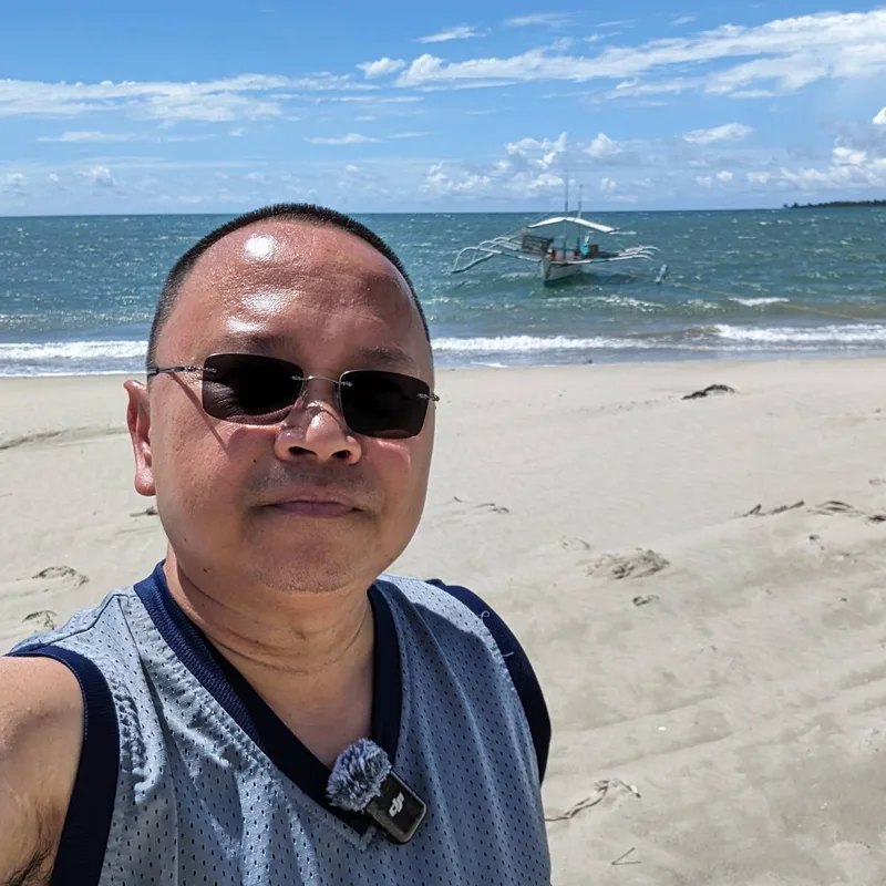 Man in sunglasses and tank top taking a selfie on a sandy Palawan beach with traditional outrigger bangka boat in background