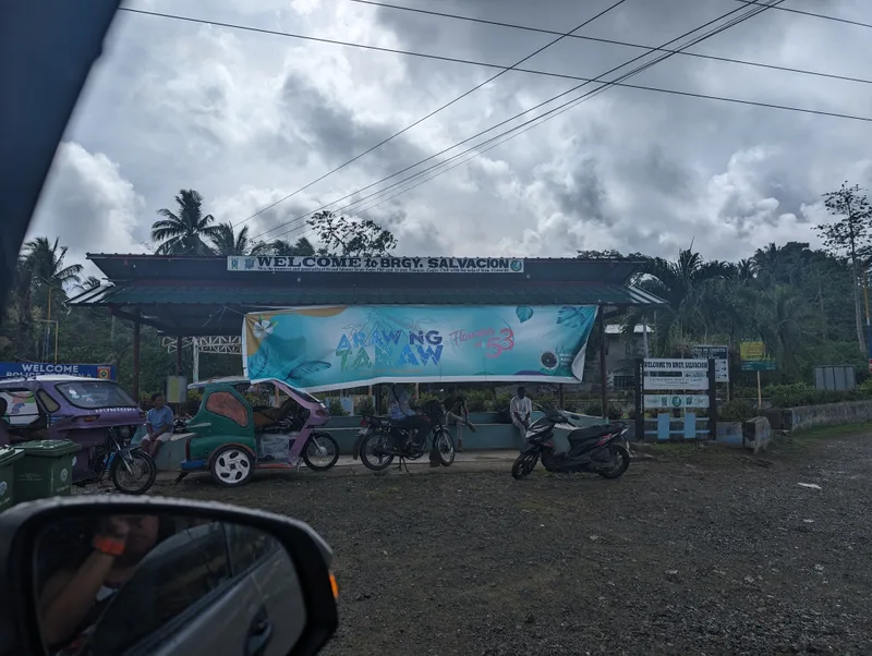 Welcome to Brgy Salvacion sign hanging over a road in Palawan, Philippines with motorcycles and tricycles parked below under an overcast sky