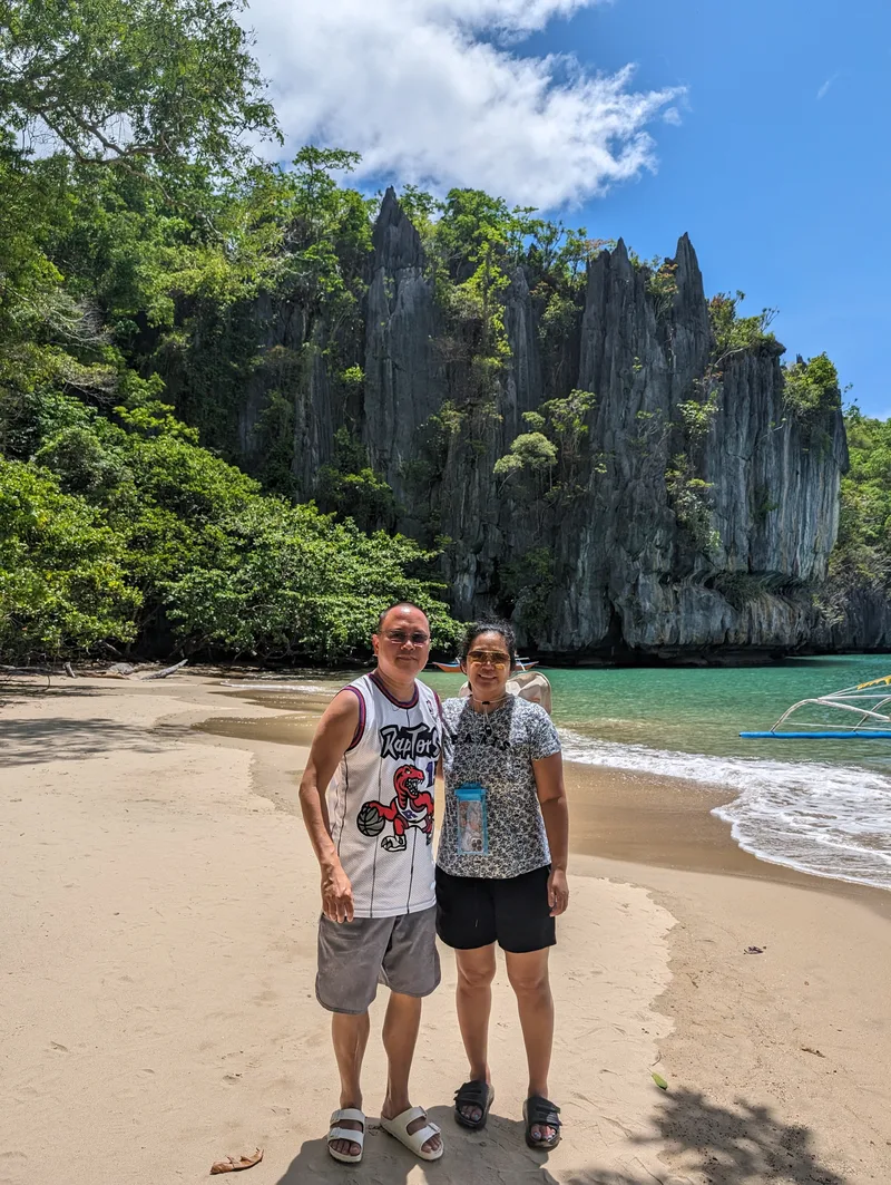 Young Filipino couple posing on a sandy beach with towering limestone cliffs covered in lush greenery behind them at the Subterranean River entrance in Sabang, Palawan, Philippines