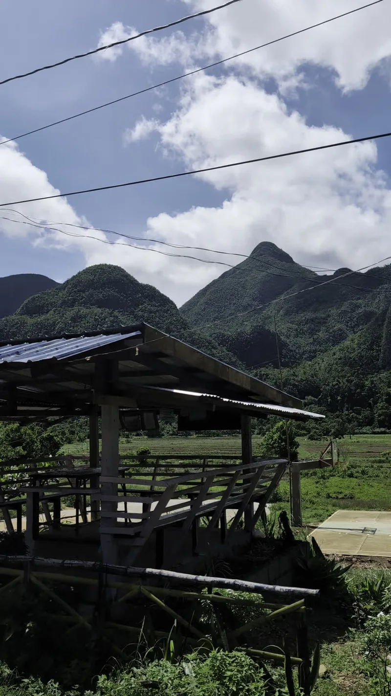 Open-air rustic shelter with a blue roof in a green field framed by lush towering mountains under a bright blue sky on the trail between the pier and the cave entrance in Puerto Princesa Subterranean River National Park, Palawan, Philippines