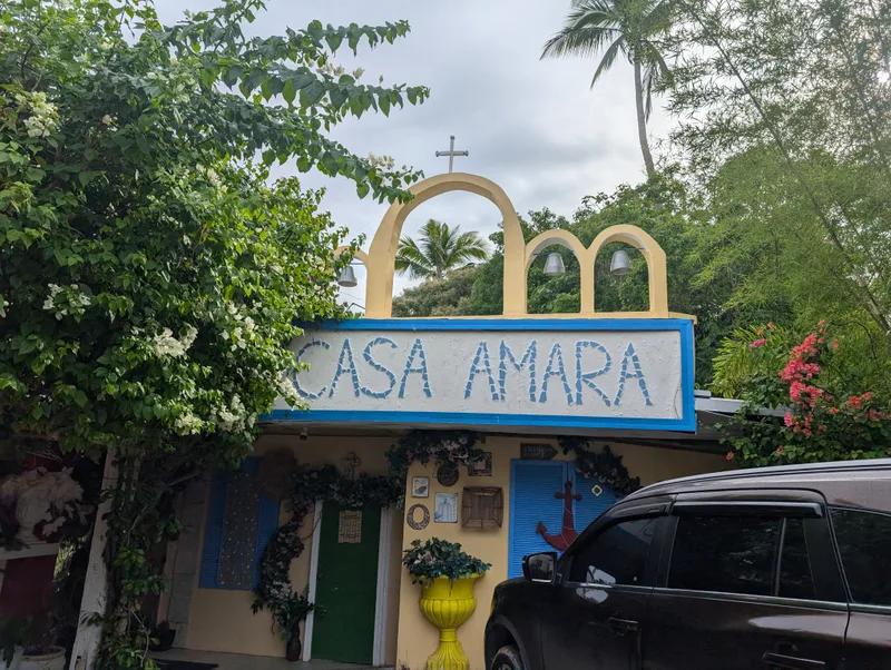 Entrance to Casa Amara — Batangas, Philippines Casa Amara resort entrance with a stone arch gate featuring bells and a cross on top, surrounded by lush green trees and flowering bushes in Batangas, Philippines.