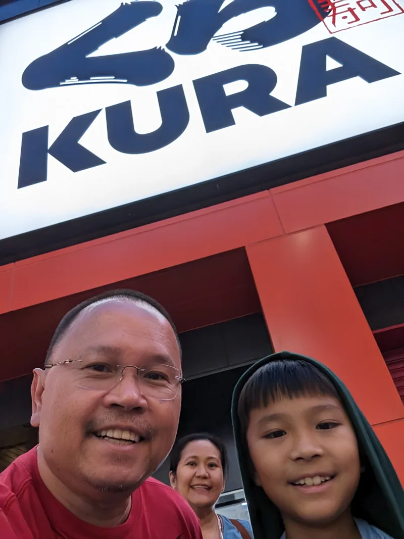 Man and boy selfie smiling in front of a large illuminated Kura Sushi sign in Osaka, Japan.