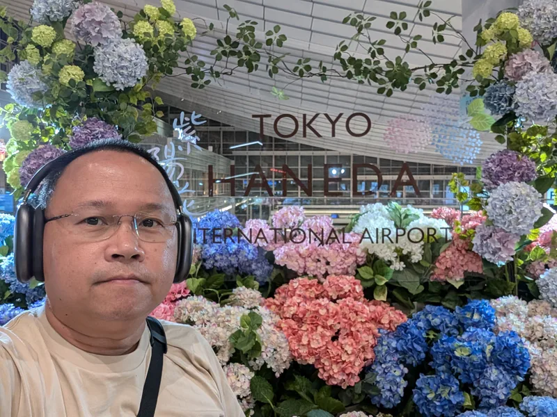 Haneda Airport, Tokyo, Japan — six hours early and nowhere to be Man wearing headphones and glasses taking a selfie in front of the Tokyo Haneda International Airport sign surrounded by a display of colorful pastel artificial hydrangeas, Tokyo, Japan.