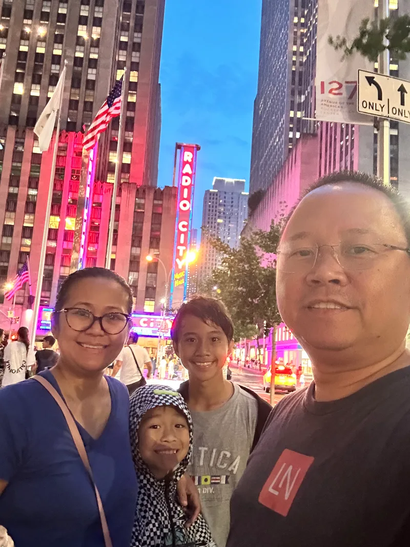Family selfie in front of Radio City Music Hall at dusk with neon lights