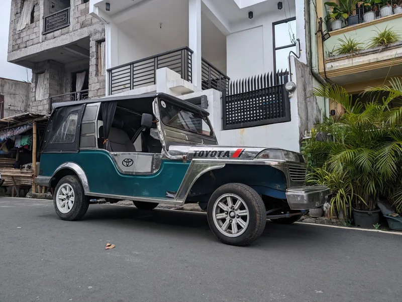 Teal and silver Owner Type Jeep parked on a street in Liliw, Laguna, Philippines in front of a modern white house with a black fence.