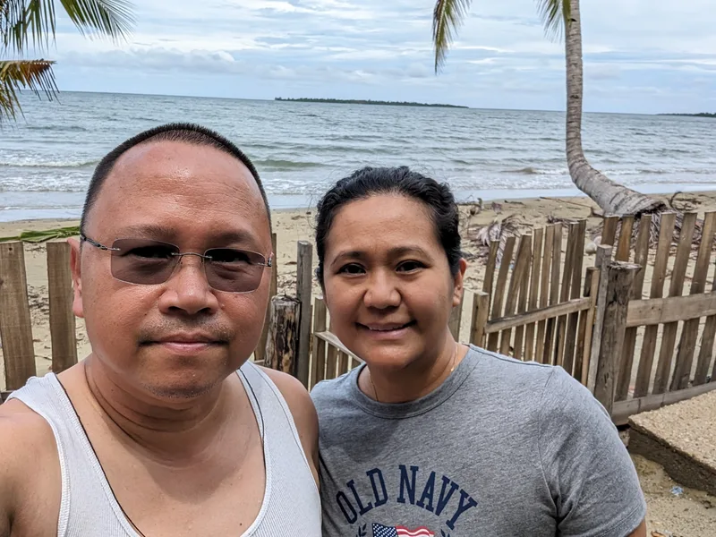 Finally some sun at the end of the day Two people smiling for a selfie on a sandy beach with a wooden fence and the ocean in the background at Henalric resort in Caramay Roxas Palawan