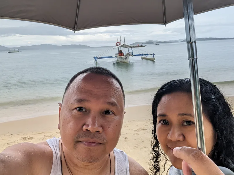 Close-up selfie of a couple under an umbrella on Port Barton beach Palawan with a traditional outrigger boat in the background