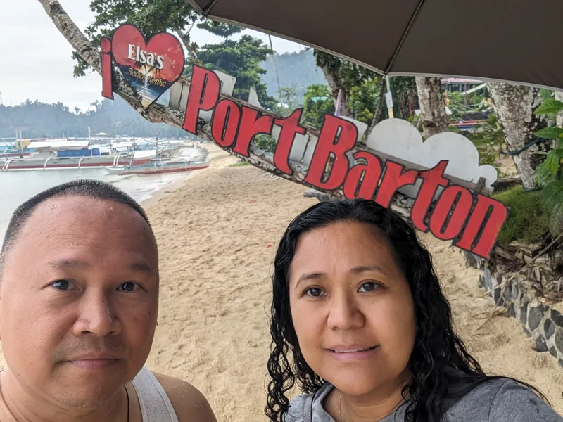 Obligatory Port Barton sign photo — no regrets Couple selfie in front of the I Love Port Barton sign on the beach with traditional outrigger boats anchored in the bay behind them