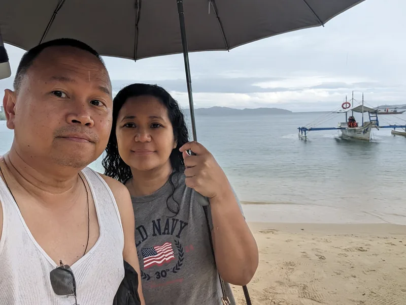 Port Barton — we got there, we saw it, we took the photo Couple selfie on Port Barton beach in Palawan Philippines with traditional outrigger boats anchored in the background on calm grey water