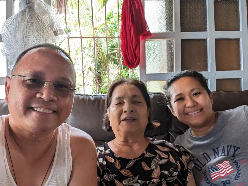 Catching up with family Selfie of man, woman, and elderly woman smiling on a couch indoors with a window and green foliage in the background at a family home in Caramay Roxas Palawan Philippines