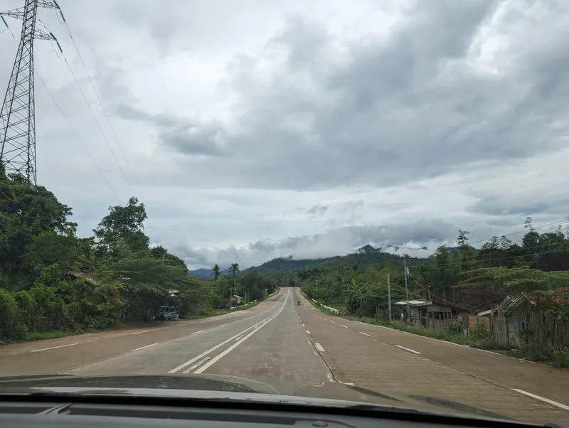 Long straight road through green rural Palawan landscape with lush trees and scattered houses and misty mountains in the background