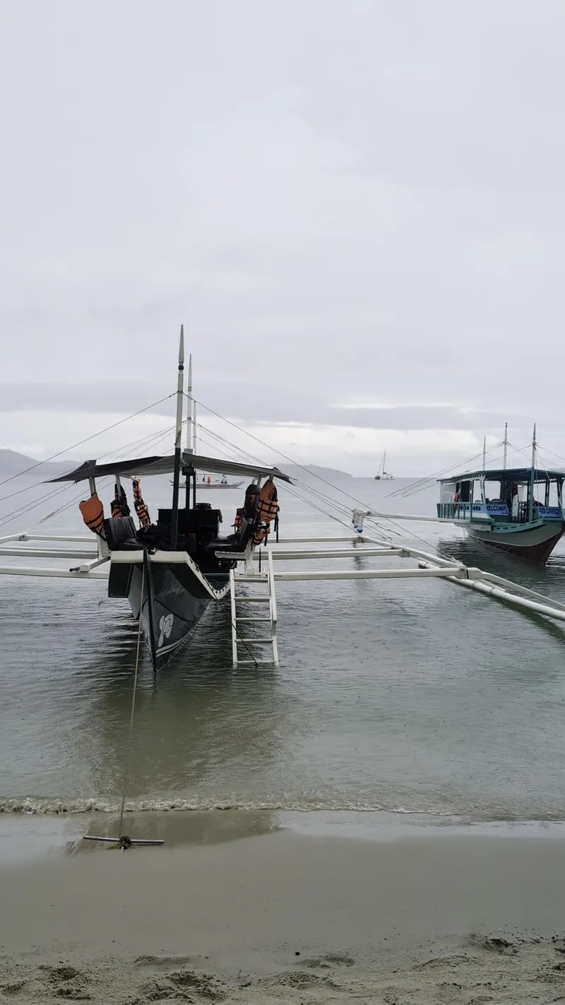 The boats waiting. Plenty of options if you're island hopping. Two traditional Filipino outrigger boats anchored in shallow calm water near Port Barton beach Palawan under an overcast sky
