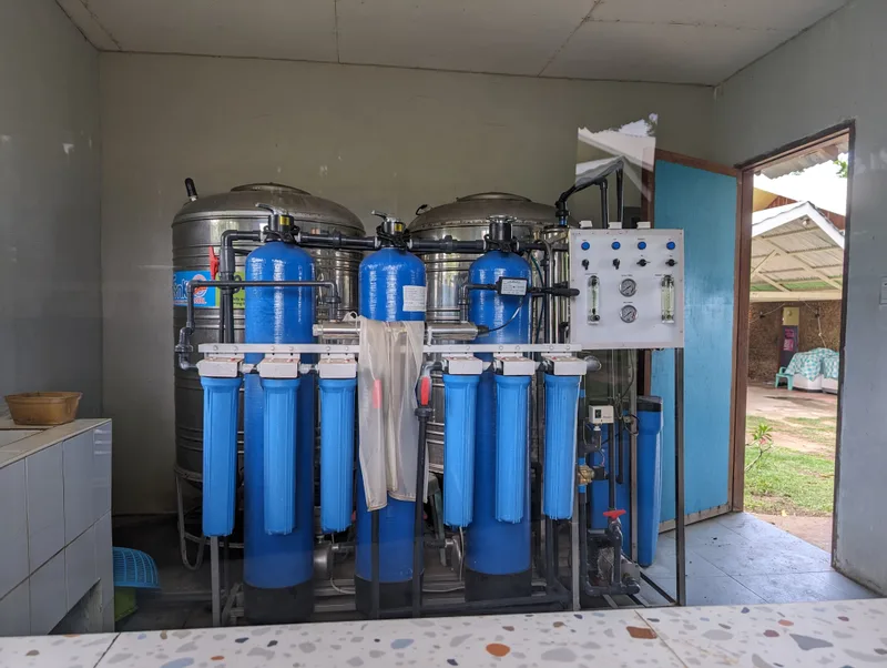 The community water station — fresh water from the well, free for the neighborhood Water purification system with large stainless steel tanks and blue filter cartridges inside an open building at Henalric resort in Palawan Philippines