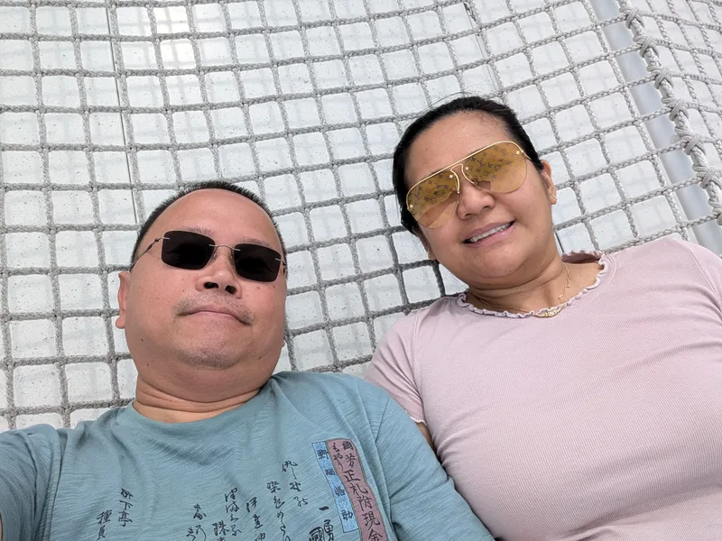 Abby and Sherwin on the Shibuya Sky rooftop net — Shibuya, Tokyo, Japan Couple lying on the white mesh net on the Shibuya Sky rooftop in Shibuya, Tokyo, Japan, smiling at the camera, man in dark sunglasses, woman in yellow aviator sunglasses.