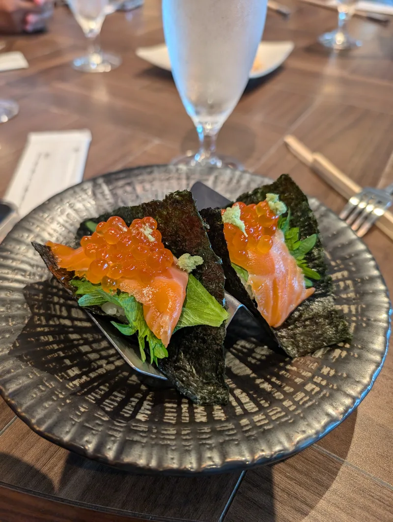 Salmon and ikura hand rolls at dinner in Shibuya, Tokyo, Japan — 7,200 yen well spent Two salmon and ikura hand rolls wrapped in nori seaweed on a dark textured plate, garnished with green leaves and wasabi, at a restaurant in Shibuya, Tokyo, Japan.