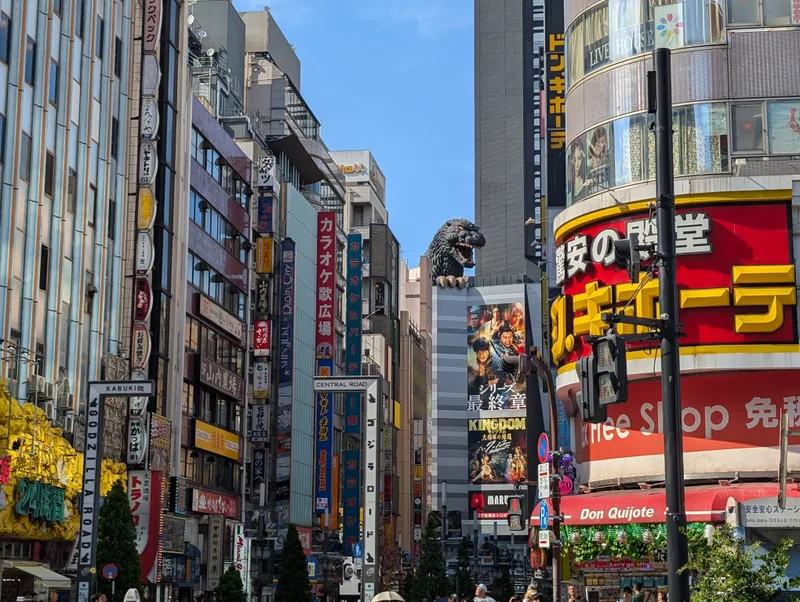 Godzilla head above the Shinjuku skyline — Tokyo, Japan Bustling Shinjuku street in Tokyo, Japan, lined with tall buildings covered in vibrant neon signs and advertisements, with the iconic Godzilla Head visible peeking above a building in the distance.