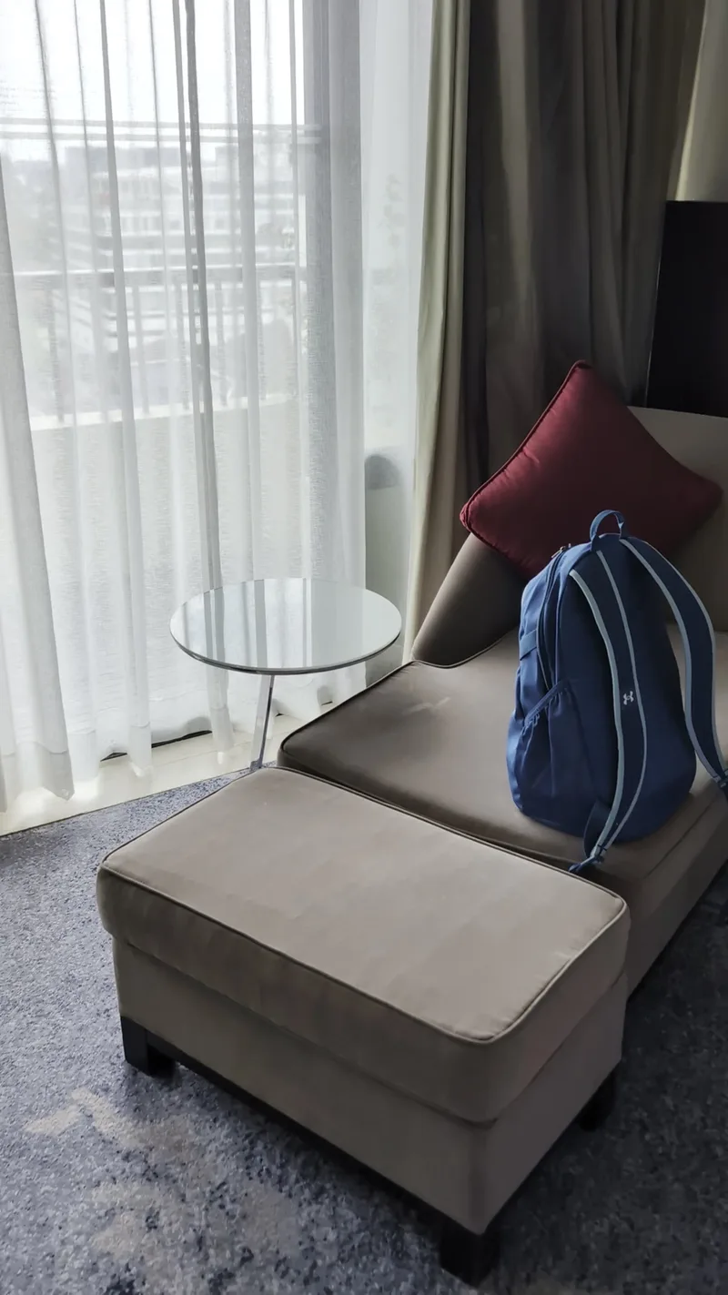 Hotel room at Sofitel Philippine Plaza with chaise lounge, red pillow, and blue backpack near sheer white curtains.